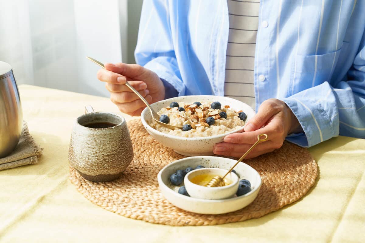 Une femme savoure un bol de céréales complètes agrémenté de baies fraîches, d'amandes croquantes et d'une touche sucrée de miel. Une manière gourmande et saine de démarrer la journée avec vitalité et bien-être.