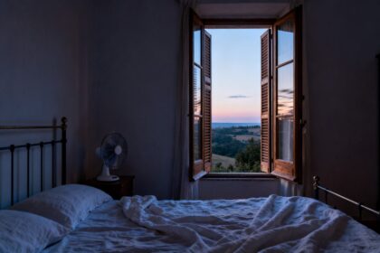 Chambre claire avec literie en lin, volets entrouverts et petit ventilateur près du lit en soirée d été.