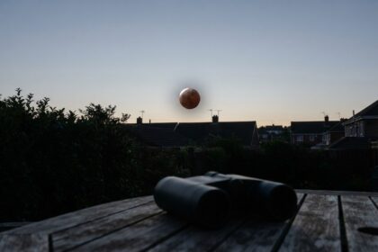 Lune cuivrée basse au-dessus de toits pavillonnaires à l'aube, jumelles posées sur une table de jardin en bois