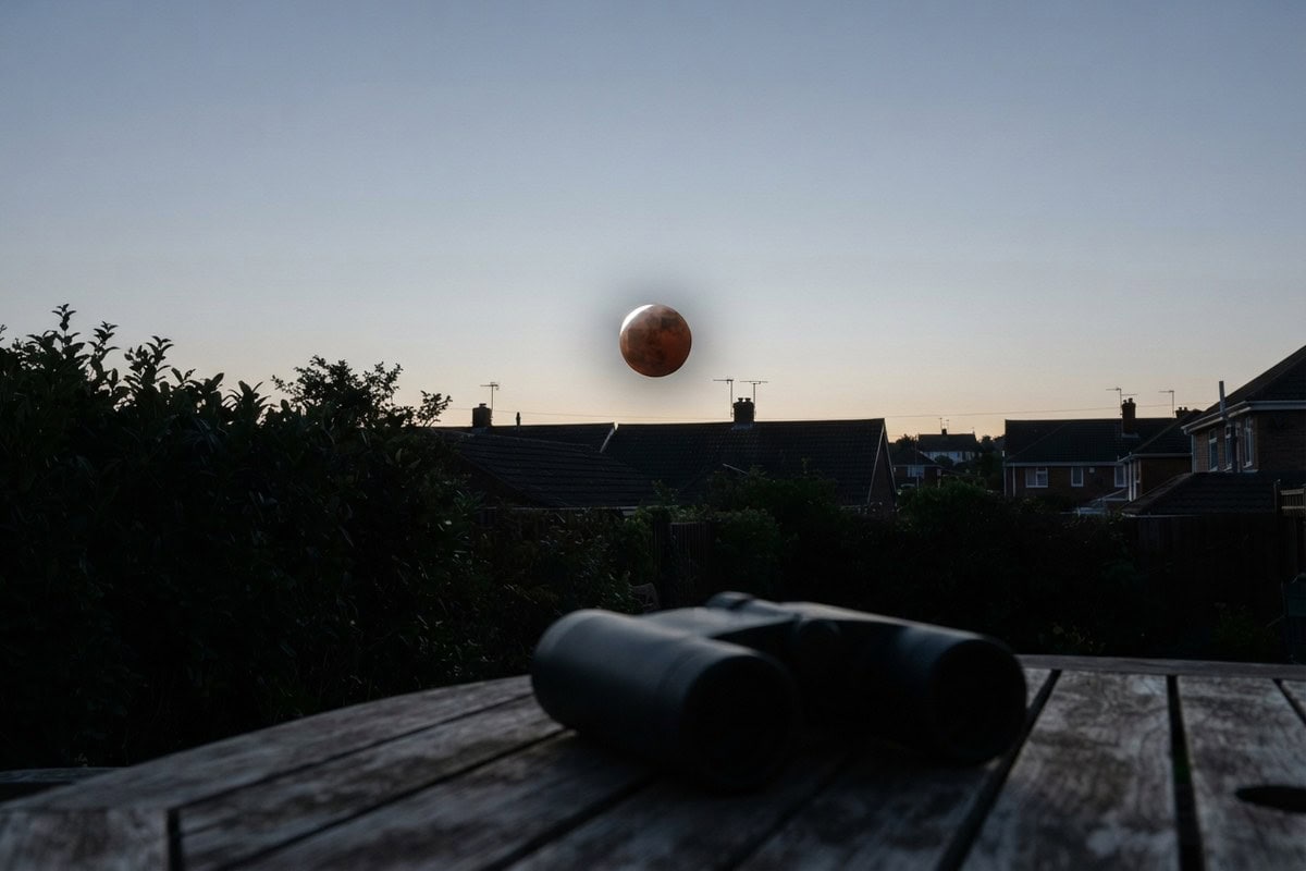 Lune cuivrée basse au-dessus de toits pavillonnaires à l'aube, jumelles posées sur une table de jardin en bois