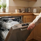 Main refermant un lave-vaisselle Bosch en inox rempli d'assiettes et de verres, dans une cuisine en bois ensoleillée
