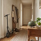Trottinette électrique noire posée dans l'entrée d'un appartement, près d'un portemanteau et d'un casque vert