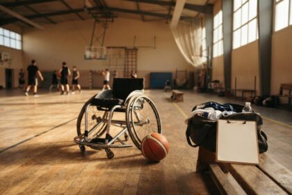 Fauteuil roulant de sport et ballon de basket au sol dans un gymnase, sac et porte-bloc posés sur un banc