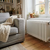 Main tournant la tête thermostatique d'un radiateur blanc près d'une fenêtre, à côté d'un canapé gris et d'un plaid