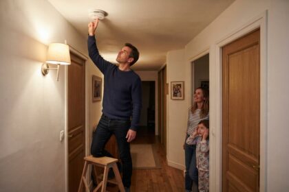 Un homme sur un escabeau teste un détecteur de fumée au plafond d'un couloir, sous le regard d'une femme et d'une fillette