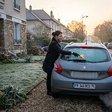 Femme grattant le givre de la lunette arrière d'une Peugeot grise, fumée d'échappement, rue au petit matin