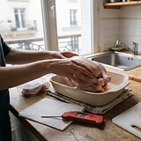 Mains déposant un poulet cru dans un plat, à côté d'un thermomètre de cuisson, d'une planche à découper et d'un évier sec