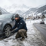 Homme accroupi posant une chaîne à neige sur la roue avant d'une voiture grise, au bord d'une route de montagne enneigée