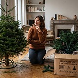 Femme agenouillée entre un sapin naturel sur son pied et un sapin artificiel encore dans son carton, dans un salon